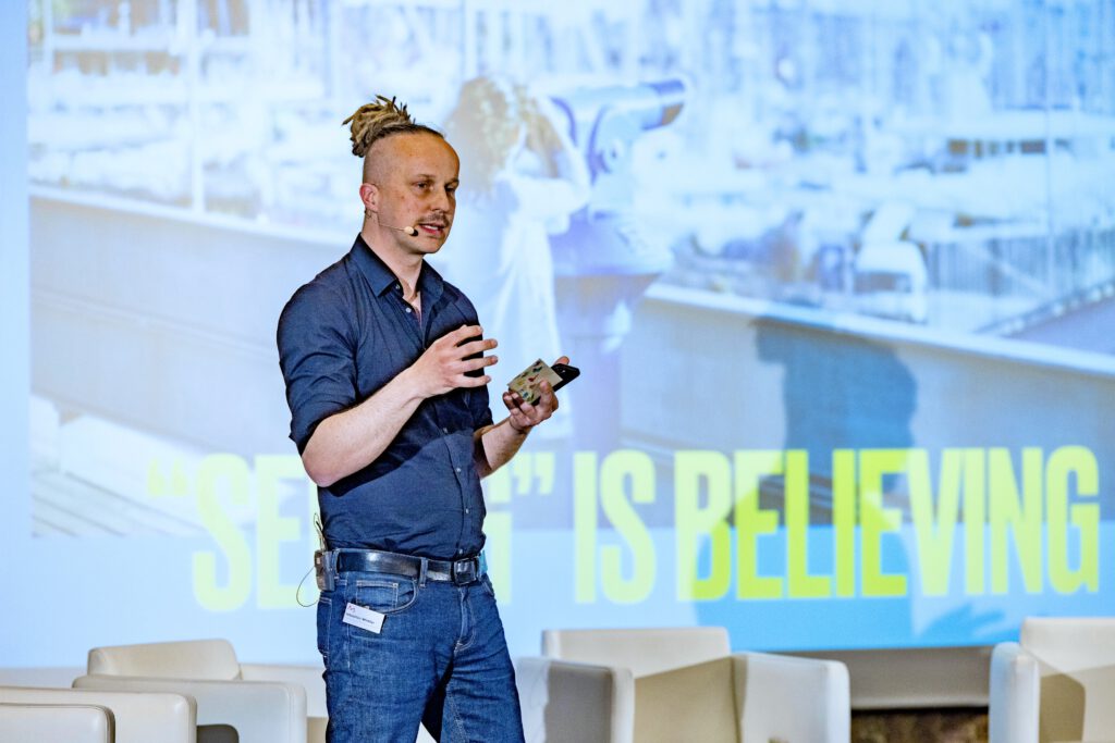 Sebastian Winkler auf der Bühne mit Smartphone in der Hand, Leinwand mit „Seeing is believing“, Mediaan 2022