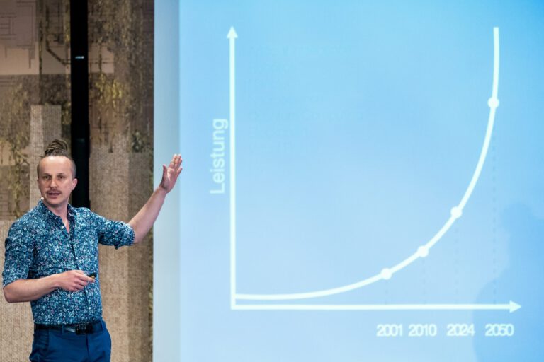 Sebastian Winkler auf der Bühne vor einer Folie mit einer exponentiell steigenden Leistungskurve von 2001 bis 2050